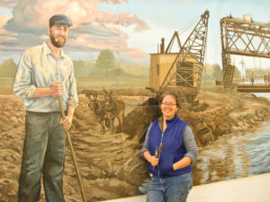 Stacy Kirby was working on affixing the mural panels when photographer Rick Nicholson captured this image of the artist and her work. The mural depicts Erie Canal reconstruction 100 years ago and is displayed on the side of a building used by the Brockport Department of Public Works at 38 East Avenue in Brockport Village.