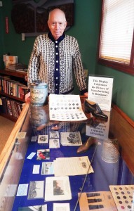At the Sweden Senior Center, Bill Andrews stands at a display case containing buttons, dolls, tin ware, fishing tackle, shoes, and other artifacts of Brockport’s manufacturing history.  A kerosene can, a woman’s shoe and a card of buttons are placed on top for better photo visibility.