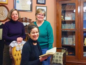 The Mary Jane Holmes room is visited by volunteers (l to r) Roberta Hesek, Sarah Hart and Maggie LaPierre. Holmes’ books in archival sleeves are organized in the cabinet in the background.  This is the first room completely recorded by PastPerfect software with every item measured and photographed.