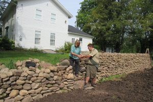 Jean Colby and Donovan Berbeneciuc place additional stones on the wall at the historic Colby/Pulver House.  All stones were gathered from fields at Colby Farms and transported to the site. Colby says the rocks are a mix of different materials including granite, Medina sandstone and shale. Jean is standing on the steps which cut into the wall and lead to the house. K. Gabalski photo 