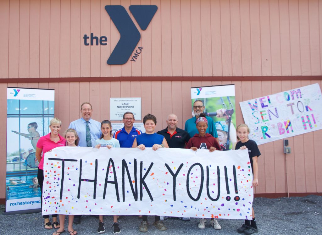 Campers presented a banner to thank those who made the upgrades at Camp Northpoint possible. Shown in the back row (l-r): Leslie Allen, Board Chair of Northwest YMCA in Greece, and owner of Grade A Flooring which installed the new turf floor at the Camp Northpoint inside the program barn; Senator Joseph E. Robach; Bill Sauers, longtime YMCA volunteer and owner of Sunday Siders which supplied the materials to make the program barn a three-season facility; Aaron Allen, co-owner of Grade A Flooring; George Romell, President and CEO of YMCA of Greater Rochester.
